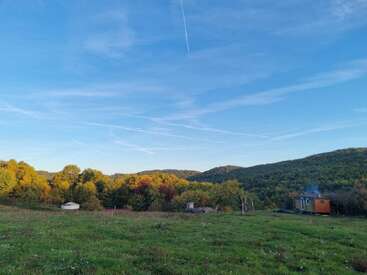 Un paisaje pintoresco con verdes colinas onduladas, árboles dispersos que lucen los colores del otoño, una pequeña construcción blanca y una casita. Cielo azul con tenues estelas de condensación en lo alto.
