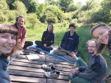 Seis personas están sentadas en una plataforma de madera al aire libre, sonrientes y relajadas. Están rodeadas de exuberante césped verde y árboles, y disfrutan juntas de un día soleado.