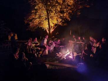 Un grupo de personas se reúne por la noche alrededor de una hoguera resplandeciente, tocando instrumentos musicales, cantando, sonriendo y disfrutando de la compañía mutua bajo un árbol iluminado.