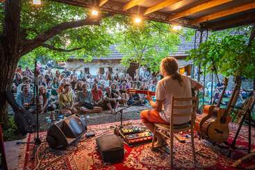 Un músico actúa en un pequeño escenario al aire libre bajo los árboles, tocando la guitarra para un público atento sentado en el suelo, creando un ambiente acogedor, íntimo y relajado.