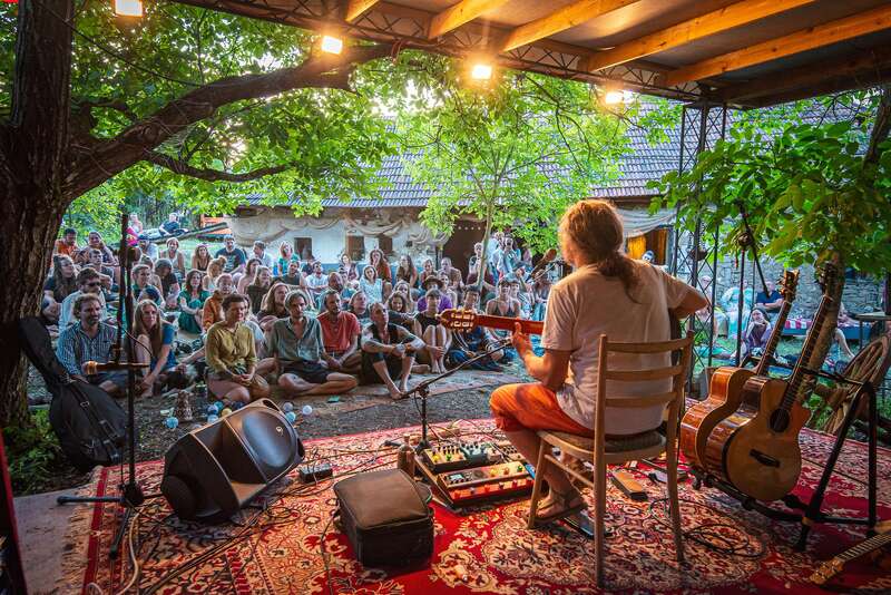 Un músico actúa en un pequeño escenario al aire libre bajo los árboles, tocando la guitarra para un público atento sentado en el suelo, creando un ambiente acogedor, íntimo y relajado.