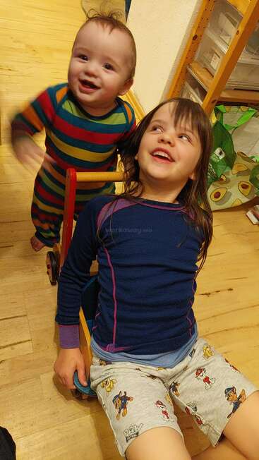 Zwei kleine Kinder spielen vergnügt in einem Haus. Das ältere Kind sitzt auf einem kleinen Wagen, während das jüngere dahinter steht. Beide lächeln und genießen den spielerischen Moment miteinander.