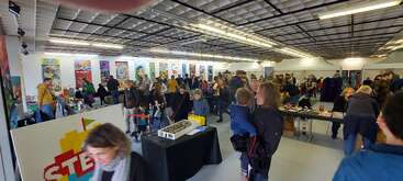 Eine geschäftige Veranstaltung in einer Halle mit vielen Menschen, die sich an Tischen und Auslagen umsehen. Bunte Poster säumen die Wände. Familien, Kinder und Erwachsene erkunden MINT- oder LEGO-Ausstellungen.
