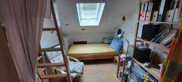 Ein gemütliches Schlafzimmer im Dachgeschoss mit einem Dachfenster, einem Holzbett, blauen Kissen, einem Lesesessel, einer Leiter, Bücherregalen, Ordnern, kleinen Lampen und einer Vielzahl von Büchern.