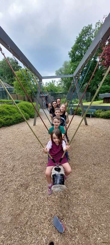The image shows a group of children and an adult on a swing set in a park, with the children sitting on a large swing.