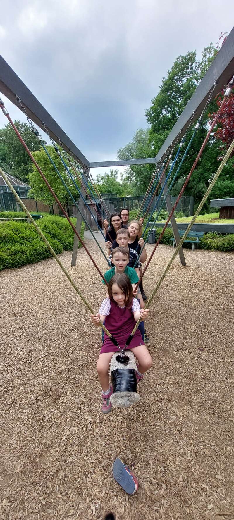 Das Bild zeigt eine Gruppe von Kindern und einen Erwachsenen auf einer Schaukel in einem Park, wobei die Kinder auf einer großen Schaukel sitzen.