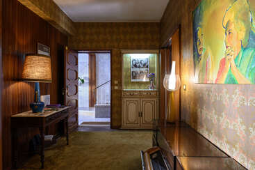 Pasillo vintage con papel pintado estampado, alfombra y muebles de madera. Gran retrato colorido, lámpara de mesa, armario, fotos de familia y una puerta abierta que conduce a las escaleras. Iluminación cálida.