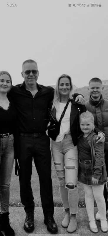 A imagem mostra uma foto de família com um homem, uma mulher e duas crianças posando para a câmera, em um fundo liso. A foto parece ter sido tirada em um ambiente interno.