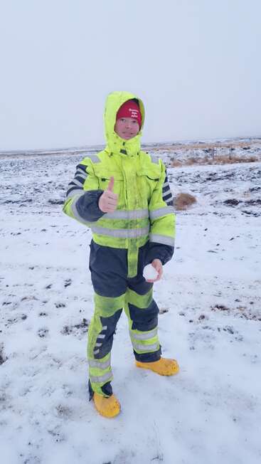 A imagem mostra uma pessoa em pé em um campo nevado, vestindo uma roupa de neve amarela e preta neon, dando um sinal de positivo e segurando uma bola de neve.