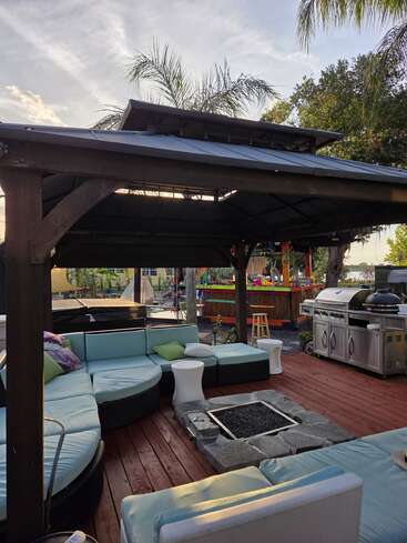 Une terrasse extérieure chaleureuse, équipée de sièges rembourrés, d'un foyer en pierre et d'un barbecue, le tout sous un kiosque en bois. Des palmiers et un coin bar festif à proximité.
