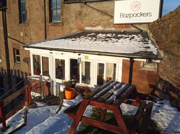 Vor dem Bazpackers Hostel gibt's einen schneebedeckten Sitzbereich im Freien mit einem Picknicktisch. Das Sonnenlicht beleuchtet das rustikale Steingebäude und die orangefarbenen Blumentöpfe.