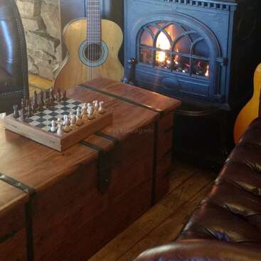 Una acogedora habitación con sillones de cuero, un baúl de madera con un tablero de ajedrez, una guitarra y una cálida chimenea, que crean un ambiente rústico y acogedor.