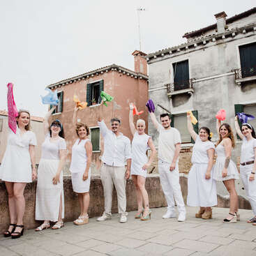 The image depicts a group of nine people, dressed in white, standing in front of two buildings, with their arms raised, holding colorful scarves.
