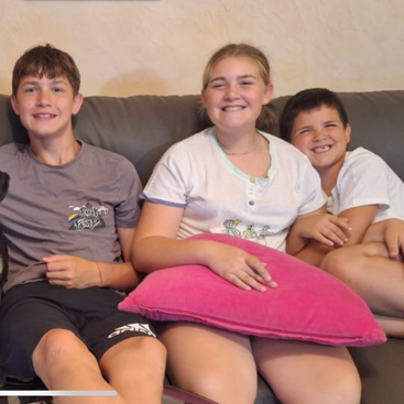 Three smiling kids sit closely together on a grey couch, holding a bright pink pillow, appearing happy and comfortable in a cozy indoor setting.