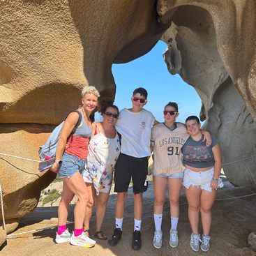Five people pose together, smiling under a large rock formation. Bright sunlight, casual summer clothes, and blue sky create a cheerful, adventurous atmosphere during their outdoor trip.
