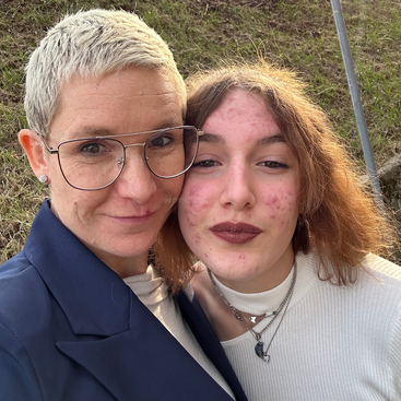 A woman with short blond hair and glasses stands beside a younger woman with curly hair, both smiling outdoors, enjoying a sunny day together.