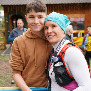 A smiling woman wearing athletic gear and a blue headwrap stands closely with a boy in a brown hoodie. They look happy, surrounded by people outdoors.