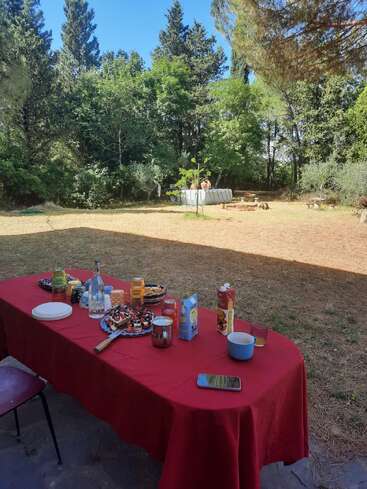 Uma toalha de mesa vermelha cobre uma mesa externa preparada para o café da manhã, cercada por árvores. Ao fundo, duas pessoas relaxam em uma pequena piscina acima do solo.