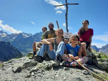 A family of five poses happily on a rocky mountain peak under a clear blue sky, beside a wooden cross and sign, surrounded by stunning scenery.