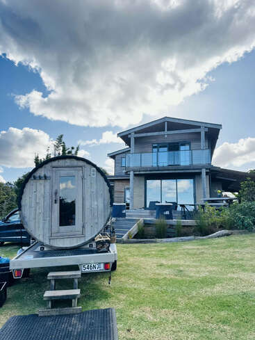 Une maison moderne en bois avec de grandes fenêtres se trouve sous un ciel nuageux. Au premier plan, on aperçoit une structure en bois en forme de tonneau sur une remorque, garée sur l'herbe.