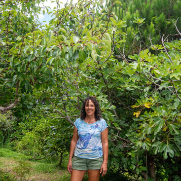 Une femme aux cheveux bouclés se tient debout, souriante, devant des arbres verdoyants. Elle porte un t-shirt bleu et un short vert, entourée d'un feuillage luxuriant et coloré.