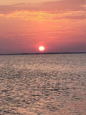 Ein atemberaubender Sonnenuntergang mit einer glühend orangefarbenen Sonne, die unter den Horizont taucht und warme Farben über ein ruhiges, sanft plätscherndes Gewässer und einen bewölkten Himmel wirft.