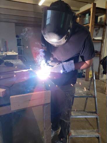 Eine Person mit Schutzvisier und Handschuhen schweißt Metallteile auf einer Werkbank und erzeugt dabei helle Funken und intensives Licht in einer Werkstatt mit Regalen.