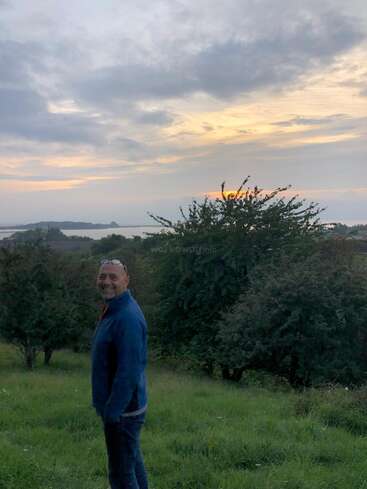 Ein Mann steht auf einer grasbewachsenen Wiese und lächelt in die Kamera. Hinter ihm sind grüne Büsche, entferntes Wasser und ein bunter, wolkenverhangener Sonnenuntergangshimmel zu sehen.