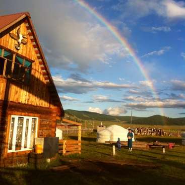Das Bild zeigt eine heitere Szene mit einem Holzhaus, einer Jurte und einer Person, die an einem teilweise bewölkten Tag auf einem Feld unter einem leuchtenden Regenbogen steht.