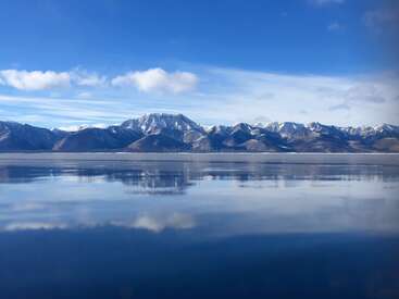 Une chaîne de montagnes sereine, couronnée de neige, se reflète parfaitement dans les eaux calmes en contrebas. Le ciel bleu au-dessus renforce cette impression de tranquillité et de paix.