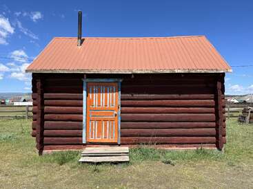 L'image représente une petite cabane rustique à l'extérieur marron, avec une porte orange et un toit orange, sur fond d'herbe sereine et de ciel bleu.