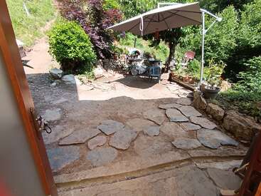 Um jardim ensolarado com degraus de pedra, arbustos verdes exuberantes, vasos de plantas, um grande guarda-sol, uma cadeira azul e um ambiente externo rústico visto de uma porta aberta.