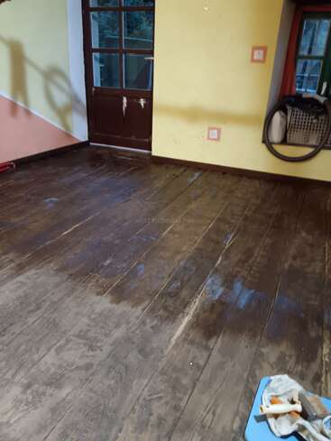Uma sala com piso de madeira velho, parcialmente limpo ou manchado, paredes amarelas e pêssego, porta marrom, janela, mangueira de aspirador de pó e materiais de limpeza à direita.