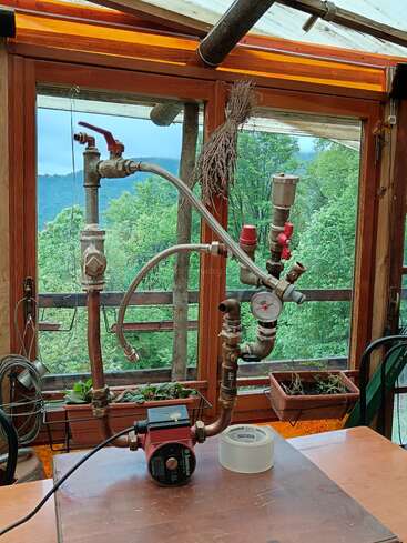 Esta imagem mostra um aparelho de encanamento caseiro com tubos, válvulas, um medidor de pressão e uma bomba, colocado em um ambiente interno com grandes janelas com vista para uma paisagem verde e florestal.