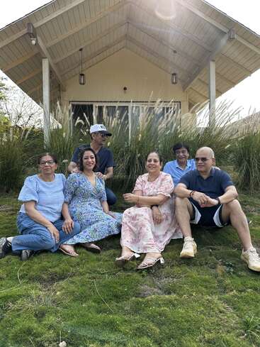 Six adults sit together on grass in front of a cozy house, smiling and enjoying a sunny day. Casual attire and relaxed moods highlight their camaraderie.