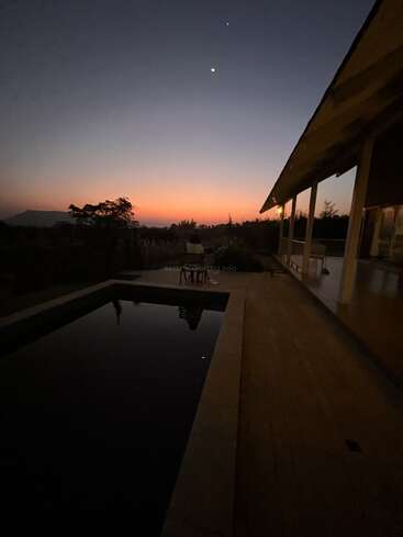 A serene dusk scene with a crescent moon, twinkling star, tranquil pool, silhouetted trees, glowing horizon, and a modern house's patio in view.