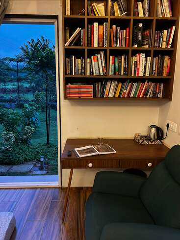 A cozy reading nook features a wooden desk, green armchair, bookshelf filled with books, and an open window overlooking a lush, green garden at dusk.
