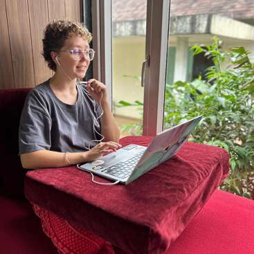 A person with short curly hair is sitting by a window, wearing glasses and earphones, using a laptop on a red cushion, smiling during a video call.