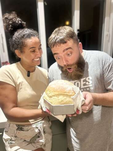Eine lächelnde Frau und ein staunender Mann bewundern glücklich einen frisch gebackenen Laib Brot in einer Pfanne und teilen einen freudigen Moment in einer Küche.