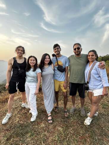 Sechs Freunde stehen im Freien zusammen und lächeln in die Kamera. Sie tragen legere Sommerkleidung und genießen eine malerische, sonnenbeschienene Landschaft mit einem teilweise bewölkten Himmel.