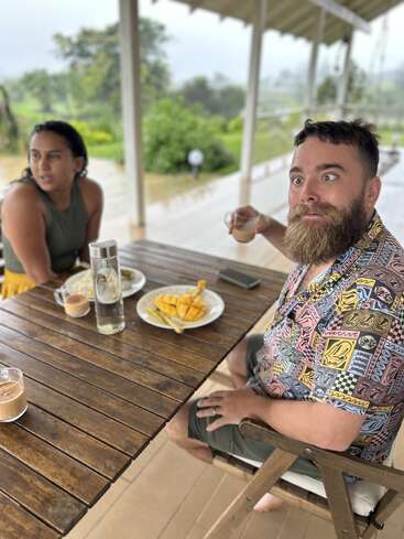 Ein bärtiger Mann mit einem überraschten Gesichtsausdruck sitzt mit einer Frau an einem Holztisch und genießt Getränke und Essen im Freien auf einer überdachten Veranda in der Natur.