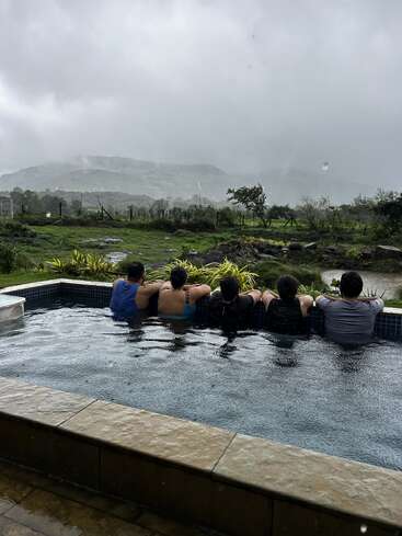 Fünf Menschen entspannen sich in einem Pool, mit dem Rücken zur Kamera, mit Blick auf eine üppige grüne Landschaft und neblige Hügel unter einem bewölkten, regnerischen Himmel. Gelassene Atmosphäre.