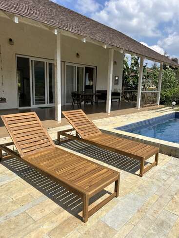 Dieses Bild zeigt zwei hölzerne Liegestühle neben einem blauen Pool, vor einer Terrasse mit Tischen, Stühlen, weißen Säulen und einem schrägen Dach.