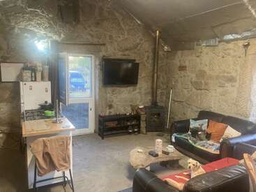 This cozy stone-walled living room features a small kitchenette, wood stove, TV, two black couches, playful dog, colorful cushions, and rustic, lived-in charm.