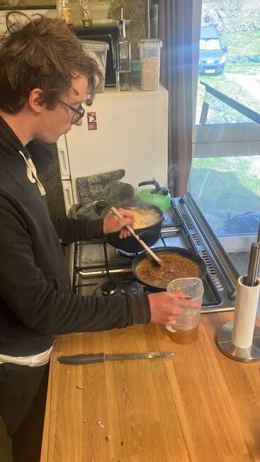A person is cooking in a rustic kitchen, stirring a pot on the stove while holding a measuring cup. Kitchen utensils and ingredients are on the counter.