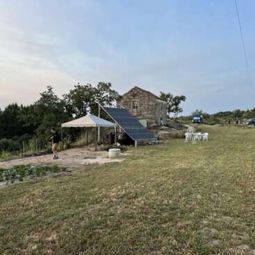 A rustic scene shows an old stone building, solar panels, white canopy, outdoor chairs, a person walking, greenery, grassy field, parked car, and trees under blue sky.