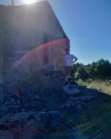 A person stands outside an old stone building, partially in sunlight. The sky is clear and blue, surrounded by trees, rocks, and grassy terrain, with visible lens flare.