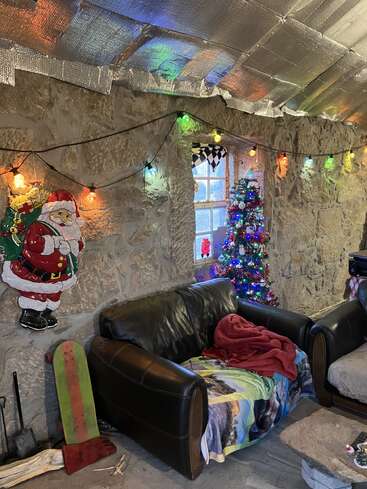 Cozy stone room decorated for Christmas, with colorful lights, a Santa wall hanging, small lit Christmas tree by the window, leather couch, and festive blankets.