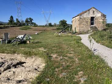 A rustic stone house stands in a rural field. Lounge chairs, power lines, green grass, a gravel path, and blue skies complete the peaceful countryside scene.
