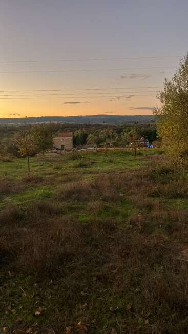 A serene countryside at sunset, a small stone building with a red roof stands among trees and grass, with hills and a glowing sky in the distance.